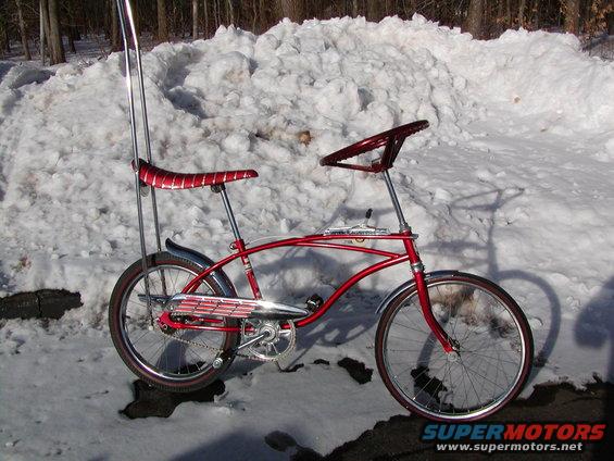 Red bicycle with banana seat, sissy bar, and steering wheel handlebars, parked in snow