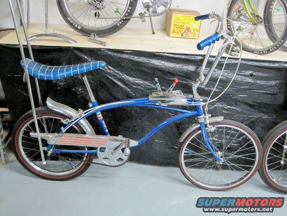 Blue bicycle with a banana seat, sissy bar, and a fake shifter on the frame. Another bicycle is partially visible to the right