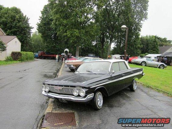 Black and white 1961 Chevrolet Impala parked on a wet asphalt road, viewed from the front three-quarter angle
