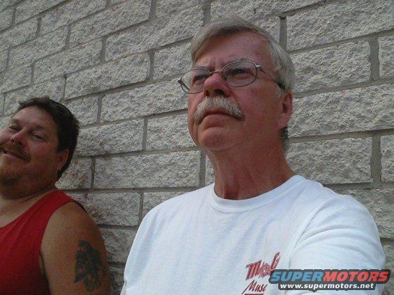 Two men, one with a mustache and glasses, the other with a red shirt and tattoo, stand against a brick wall