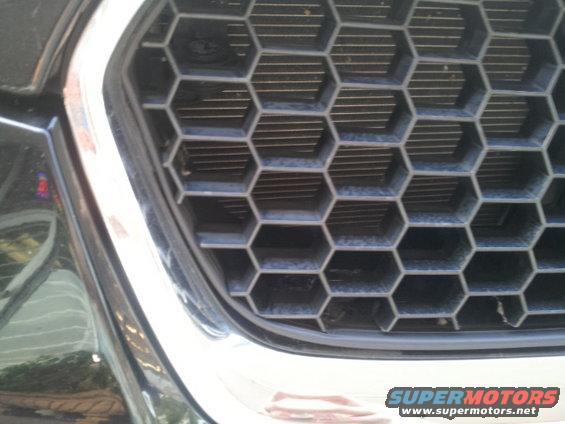 Close-up of a black vehicle's front grille with hexagonal openings and chrome trim. A 'SUPERMOTORS' watermark is visible at the bottom