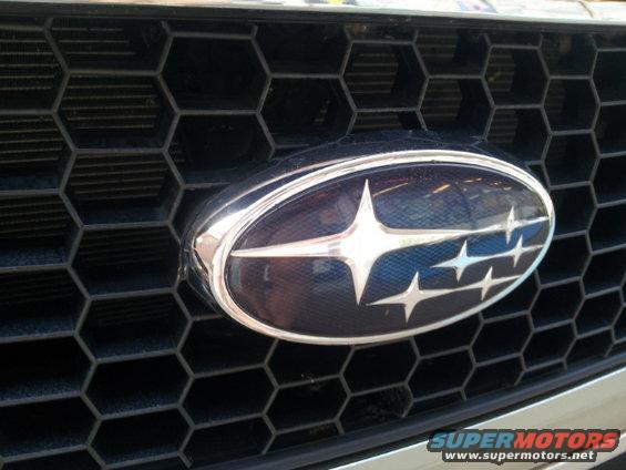 Subaru grille emblem with hexagonal pattern. Visible text 'SUPERMOTORS'