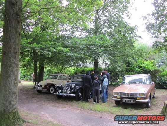 Three vintage cars parked on a dirt path under trees; two people stand near the middle car