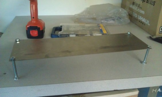 Metal shelf with four bolts for legs, next to a cordless drill and a clear plastic tool case