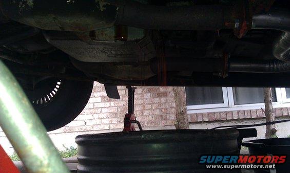 Underside of a vehicle with transmission fluid draining into a pan, with a jack visible
