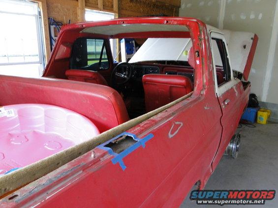 red pickup truck interior with red seats, dashboard visible. Bed has a pink inflatable pool inside. Hood is open