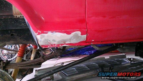 Red vehicle side panel with body filler on the lower section, showing suspension components and a wheel