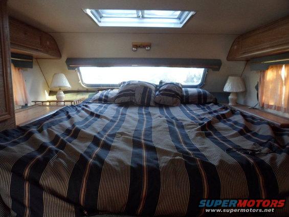 Camper interior with a large bed, striped comforter, pillows, two lamps, and a skylight