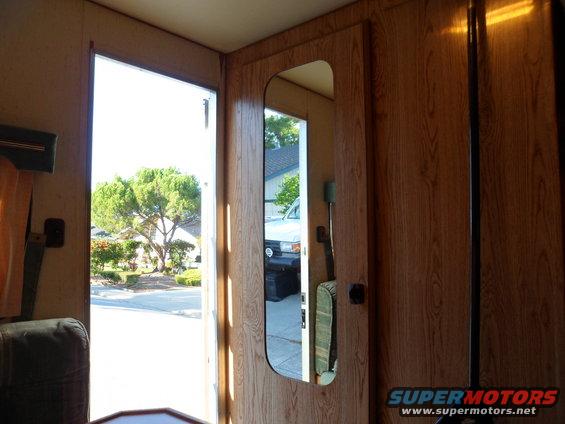 View from inside a vehicle, looking out an open door. A mirror on the wall reflects a white SUV parked outside