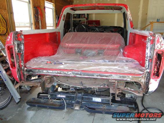 1978 Ford Bronco body, red, undergoing restoration in a workshop. Rear bumper removed, interior covered with plastic