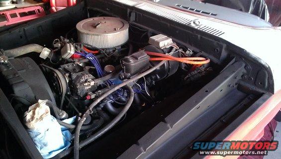 Engine bay with V8 engine, air filter, and orange spark plug wires. Blue and red hoses are visible