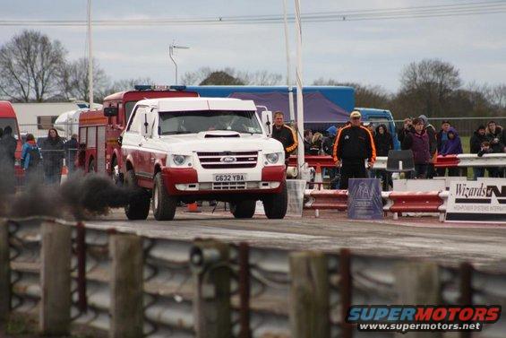 White and red SUV emitting black smoke on a drag strip, with a fire truck and spectators in the background