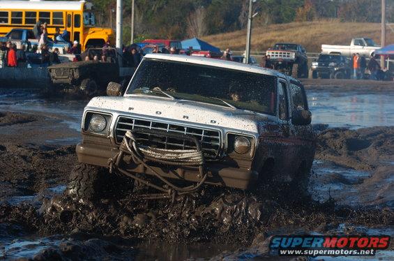 White 1978 Ford Bronco driving through mud, front grille visible, rope on bumper