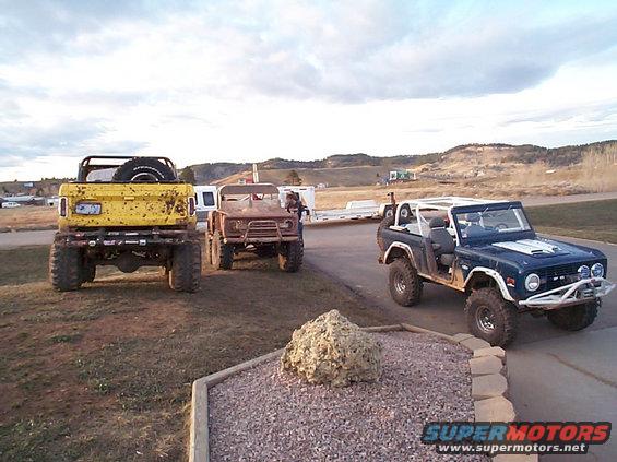 Three Ford Broncos parked on a dirt lot with hills in the background. The blue Bronco is in the foreground