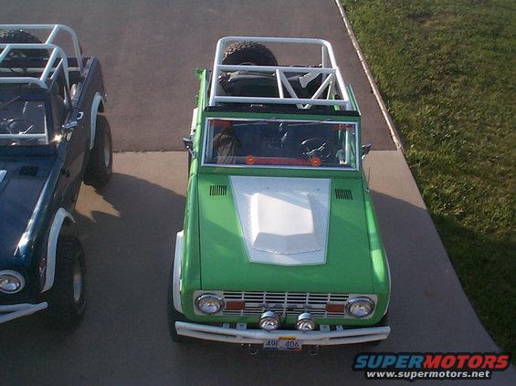 Green 1977 Ford Bronco with white hood, roll cage, and spare tire on top, parked next to a blue Bronco