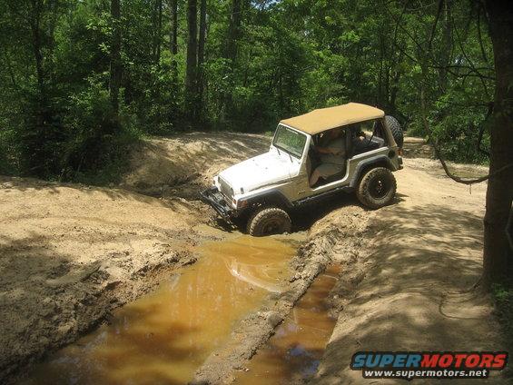 White Jeep Wrangler with tan soft top navigating a muddy trail and water obstacle