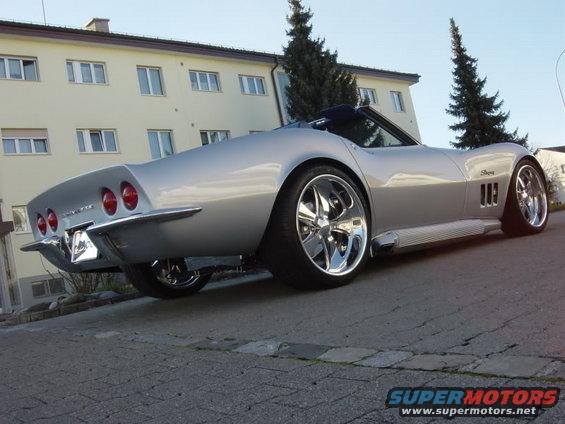 Silver convertible sports car with chrome wheels, parked on a street. Rear view shows quad taillights and side pipes