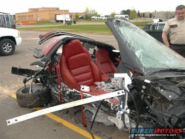 Damaged red convertible sports car with red interior, front end destroyed. A police officer stands nearby