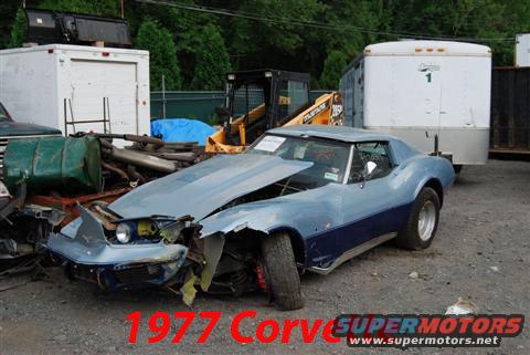 Damaged blue and light blue Chevrolet Corvette in a junkyard. Hood is up, front end is smashed