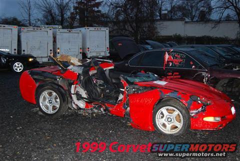 Wrecked red sports car with extensive damage to the front and side. Text overlay reads '1999 Corvette SUPERMOTORS'