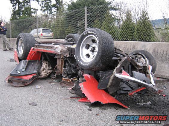 Overturned red sports car with BFGoodrich tires, showing extensive damage and a white SUV in the background