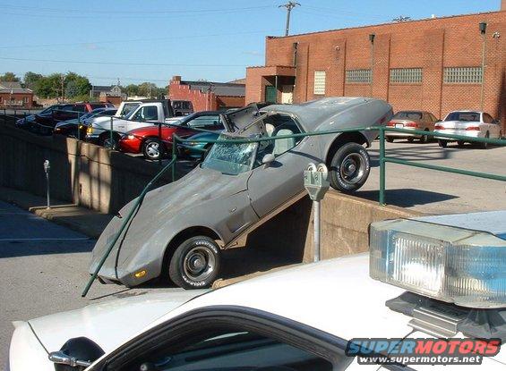 Gray Chevrolet Corvette with a damaged windshield and roof, tilted on a concrete barrier