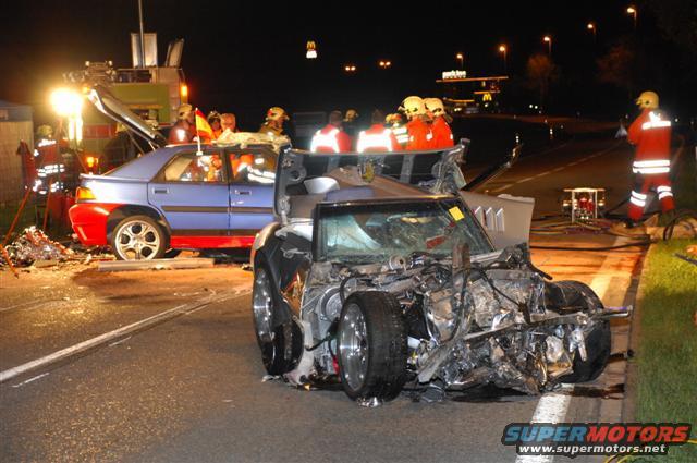 Severely damaged sports car and blue sedan involved in a nighttime crash, with emergency personnel present