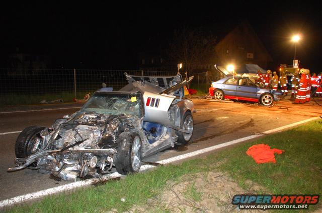 Silver sports car with front end destroyed, hood open, and driver's side door missing. Three red vents are visible on the side