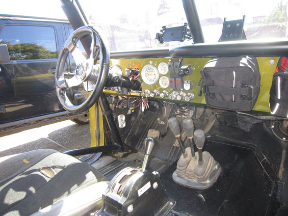 Interior view of a modified off-road vehicle with a custom dashboard, aftermarket steering wheel, and multiple gear shifters. Another vehicle is visible through the window