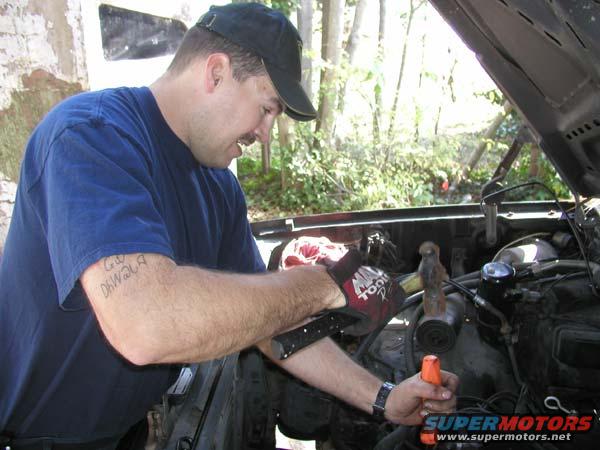 Man in blue shirt and black cap works on car engine, holding tool. Tattoo on arm