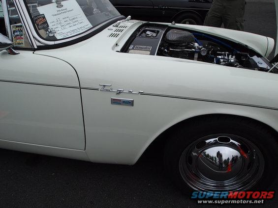 White sports car, side view, hood open showing engine, chrome hubcap, black trim