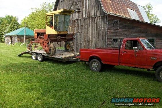 1987 Ford F-250 towing a yellow and red farm implement on a trailer next to a barn