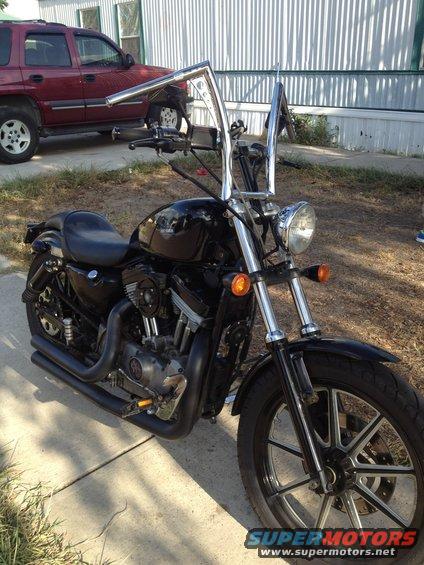 Black Harley-Davidson motorcycle with tall handlebars parked on a sidewalk. Red SUV in background