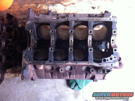 Engine block with cylinder bores and bearing caps visible, resting on a concrete floor with a wooden background