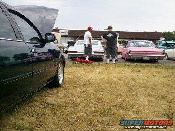 Dark sedan side view, hood open, parked at car show with other vehicles