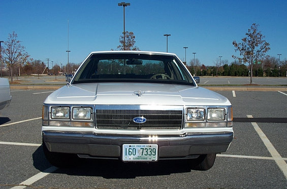 White Ford Crown Victoria sedan parked in a lot, front view. Chrome grille, rectangular headlights, and a New Hampshire license plate