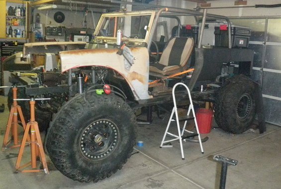 Off-road vehicle body on jack stands with large tires, roll cage, and open interior. Ladder and tools nearby
