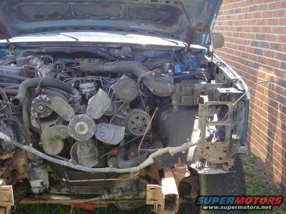 Engine bay of a blue truck, hood open, showing the engine and front chassis components. Some parts are missing or rusted