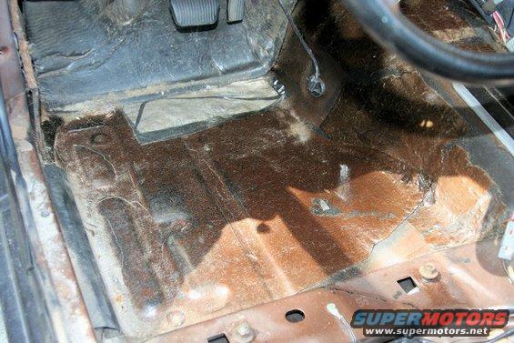 Car floor with rust and missing carpet, showing pedals and part of steering wheel