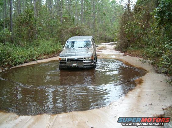 Gray Ford Bronco driving through a muddy puddle on a dirt road