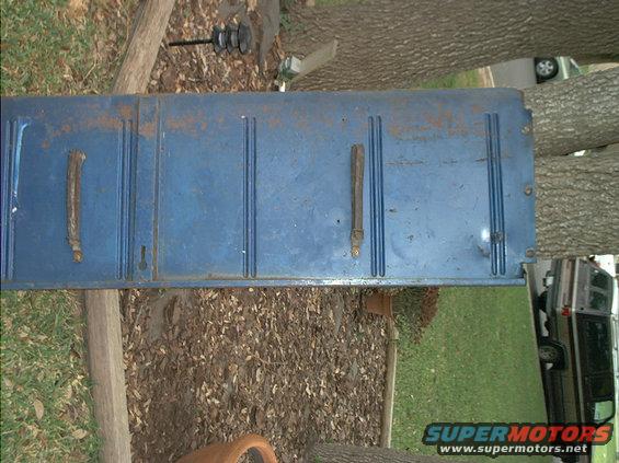 Blue tailgate with two handles and vertical lines. A vehicle is visible in the background