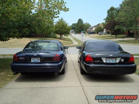 Two Ford Crown Victorias parked in a driveway, one dark blue and one black, viewed from the rear