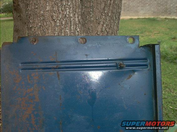 Blue metal panel with horizontal ridges, rust, and bolt holes, leaning against a tree