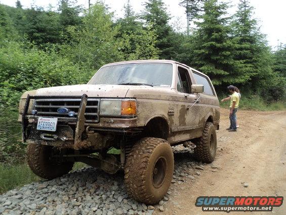 1989 Ford Bronco covered in mud, parked on a gravel and dirt road with trees in the background