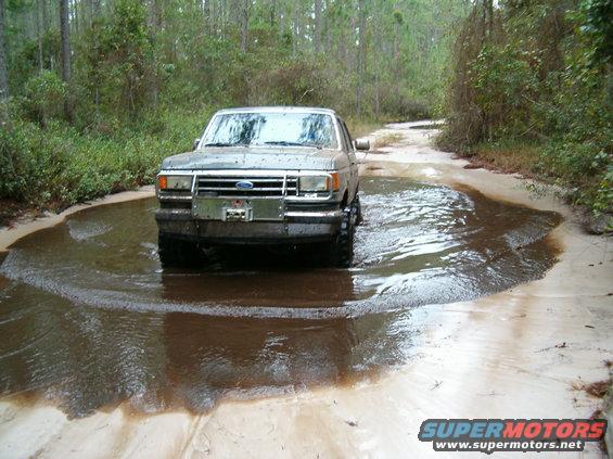 Gray Ford Bronco driving through a muddy puddle on a dirt road