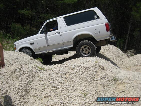 White 1992 Ford Bronco climbing a dirt mound, driver visible inside