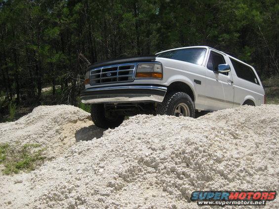 White 1994 Ford Bronco driving over a pile of light-colored dirt. Trees are in the background