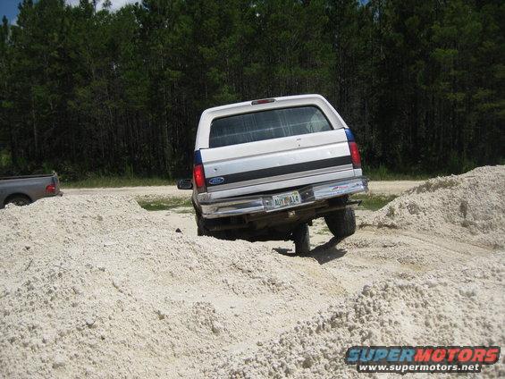 White Ford Bronco with blue stripe, rear view, driving off-road over sand mounds