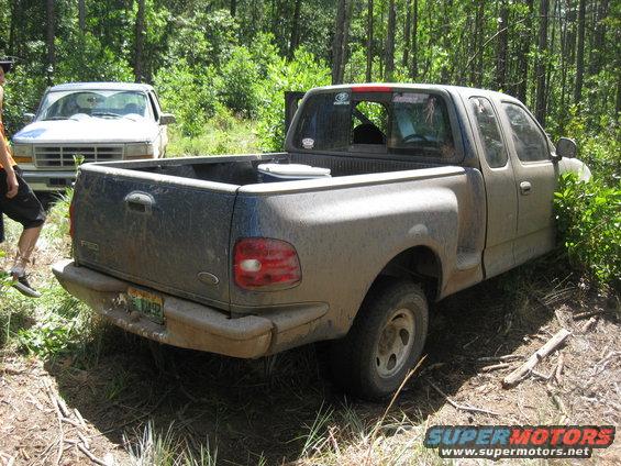Mud-covered 1997 Ford F-150 pickup truck, rear view, parked off-road with another truck behind it