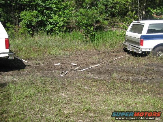 White and blue SUV stuck in mud, being pulled by another vehicle with a tow strap